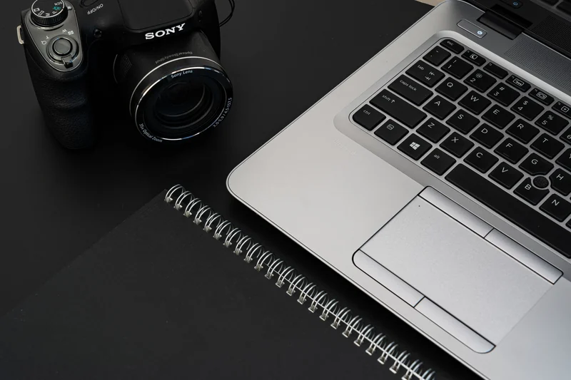 A stylish office desk featuring a laptop, camera, and spiral notebook on a dark surface.